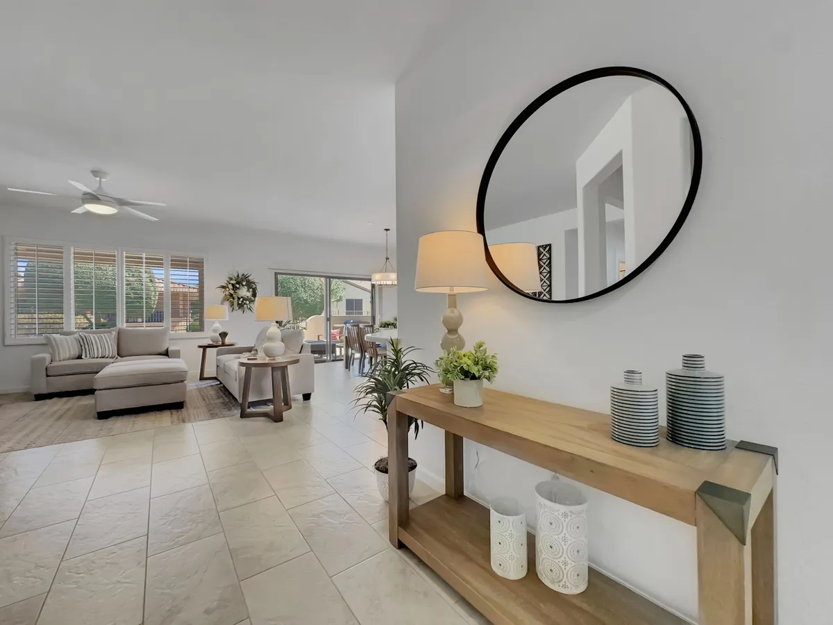 The photo showcases a welcoming living area with a wooden console table, circular mirror, green plant, and ample comfortable seating. The space is sunlit, exuding an inviting and cozy vibe suited for relaxing, entertaining, or enjoying morning coffee. The decor is tasteful, with elements that suggest both relaxation and social interaction.