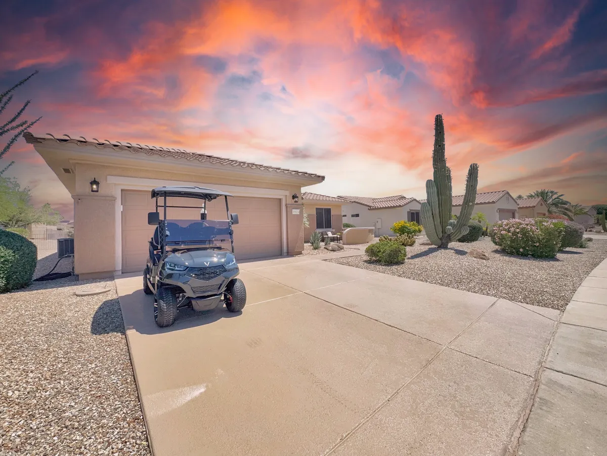 A golf cart sits in front of a driveway bordered by cacti and well-maintained landscaping, with the garage and house in the background. The scene suggests ease of access to community features and a welcoming neighborhood environment.