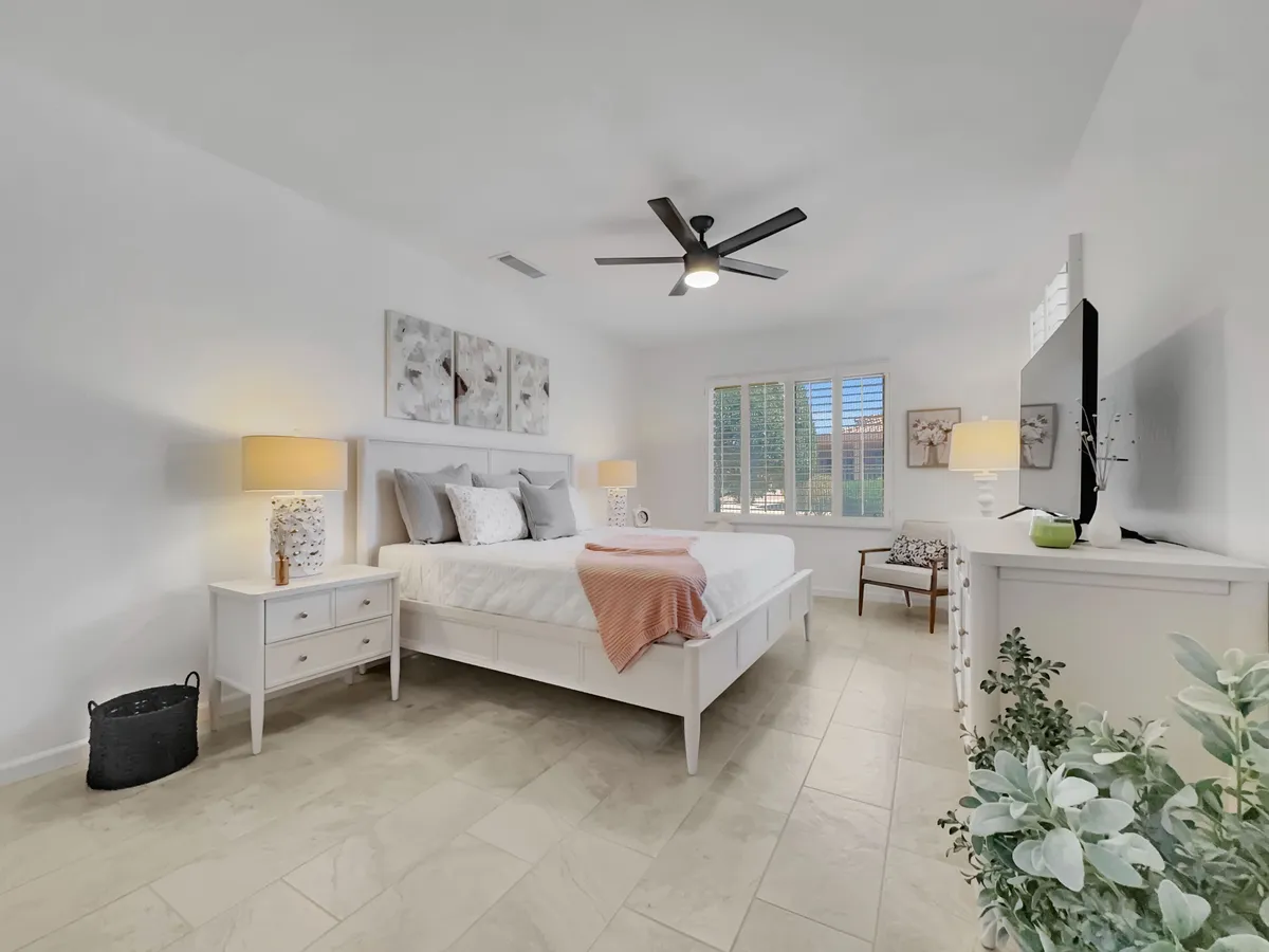 This image showcases a serene bedroom featuring a king bed dressed in crisp white bedding, flanked by nightstands with lamps, decorative wall art, a window with plantation shutters, and a plant in the corner. The space exudes tranquility and warmth, promising restful nights.