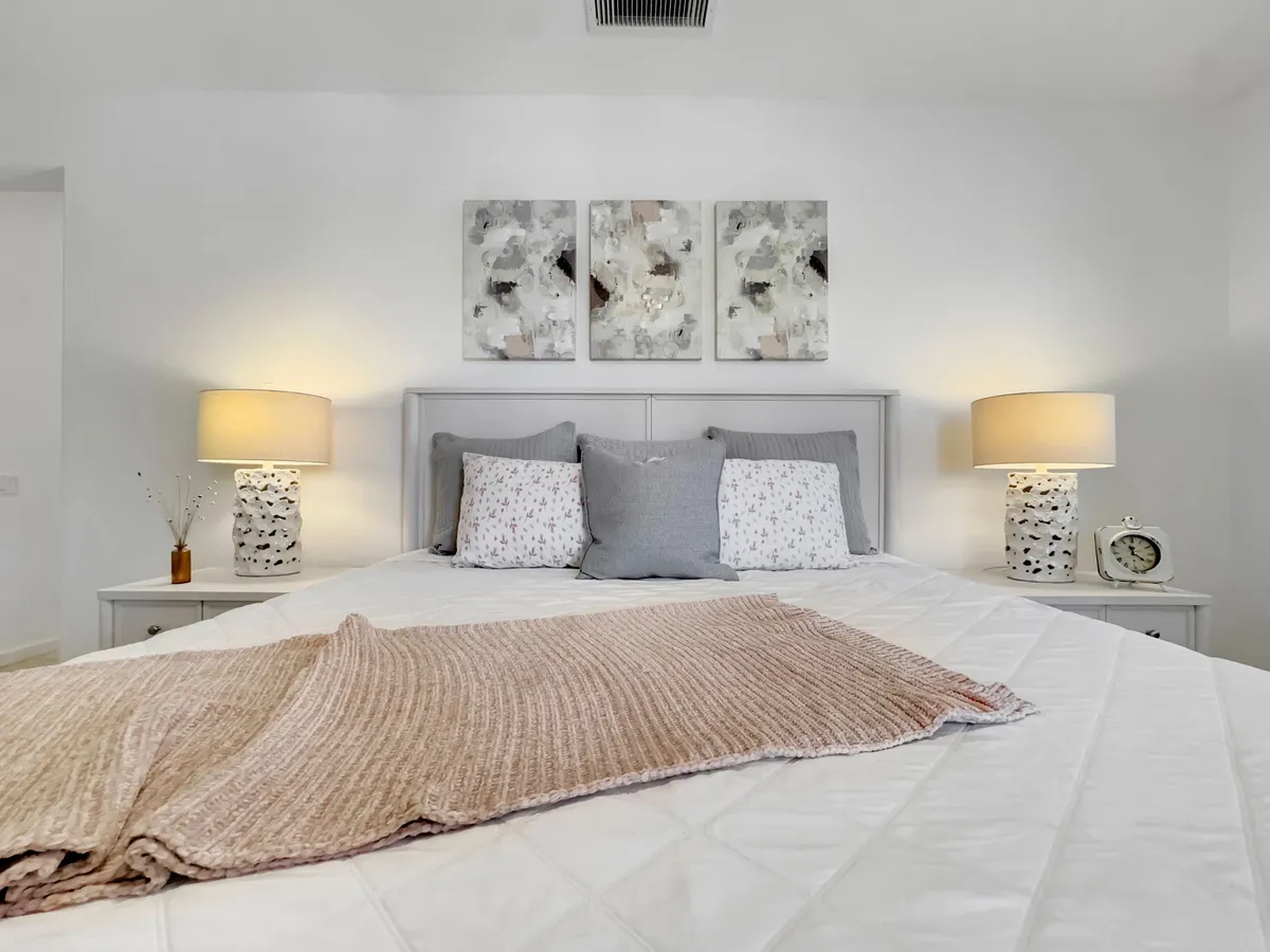 A cozy bedroom scene with a bed adorned with decorative pillows, artwork above the headboard, and soft lighting from table lamps on nightstands. The setting is intimate and inviting, ideal for relaxation.