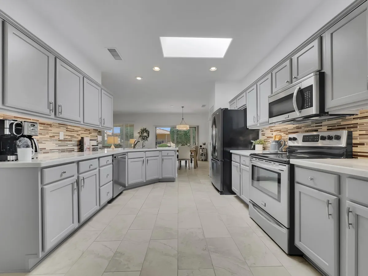 A bright, modern kitchen with a generous island at the center, featuring stainless steel appliances, a textured backsplash, and abundant natural light streaming in from a skylight. The space is clean, functional, and invites gathering for cooking or casual meals.