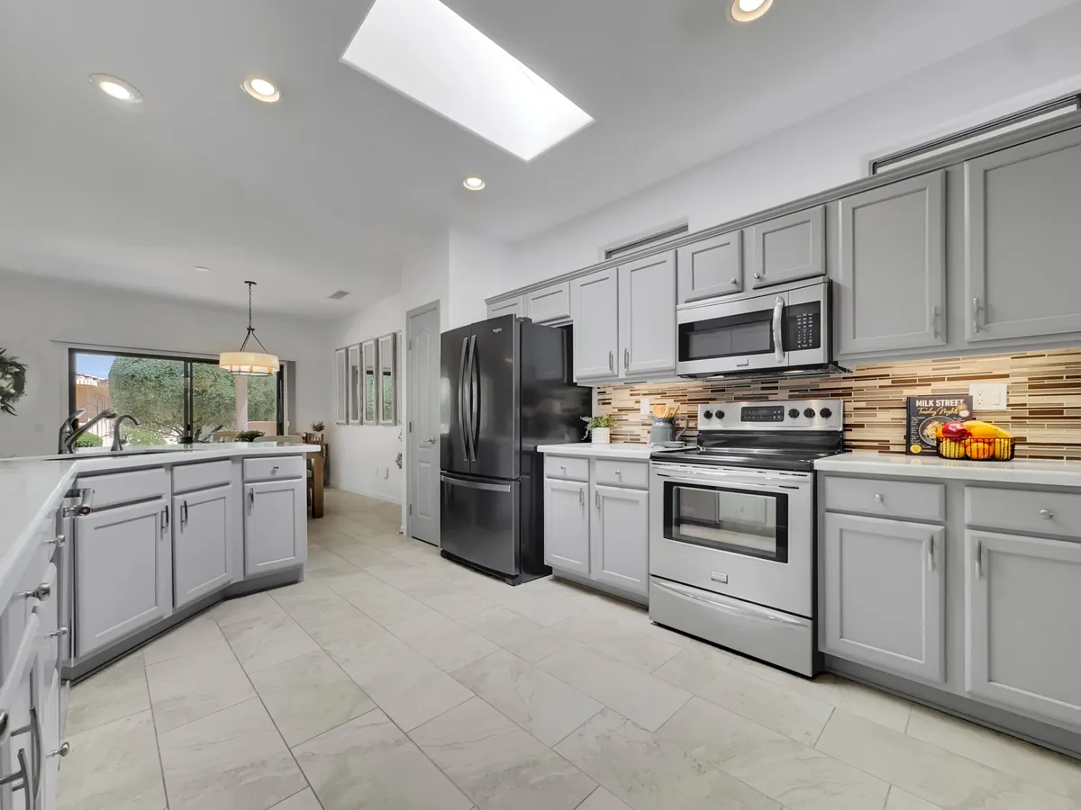 A kitchen scene with a prominent island, black refrigerator, and stainless steel appliances. There’s a window providing natural light and a warm, welcoming vibe, suggesting both practical meal prep and a space for socializing or family activities.